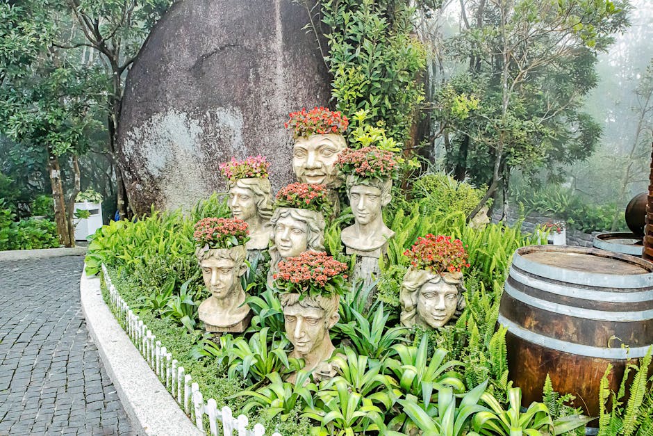 A serene garden scene featuring stone busts adorned with vibrant flowers, surrounded by lush greenery and trees.